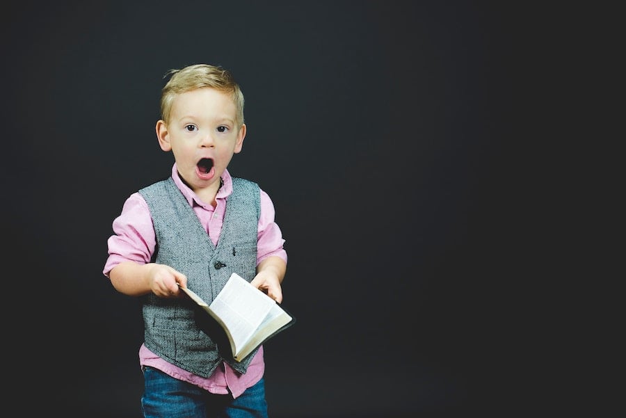 boy why a young boy stands holding a book, his mouth is wide open in shock