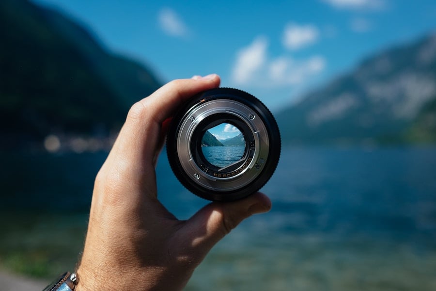 a hand holds a lens up to look at a lake; if you look through the lens, the lake is in focus, but the area surrounding is out of focus